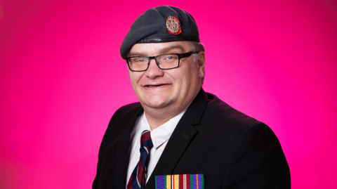 A man dressed in formal military-style attire consisting of a dark blazer with four colourful service medals, a striped tie, and a grey beret with an insignia, stands smiling into the camera against a bright pink background.