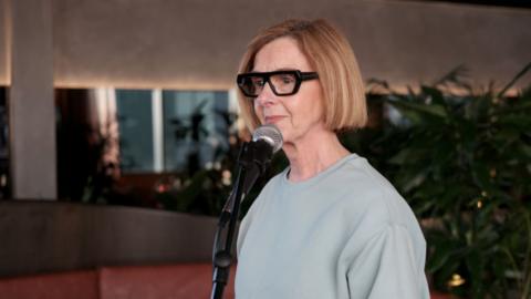 A woman with a blonde bob, black framed glasses and light green jumper is standing in front of a mic.