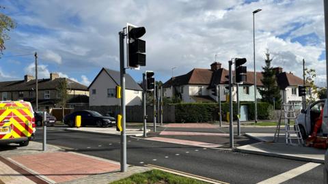 A road with cars on it with a series of traffic lights and houses and greenery in the distance.