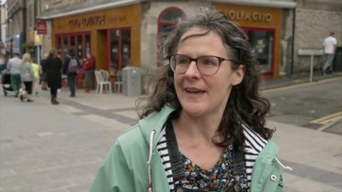 A woman standing in the street in front of a cafe. She has dark, curly hair and red glasses on.