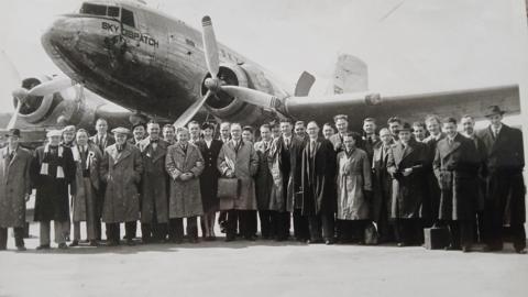 A black and white picture of about 30 men in front of an aircraft which has a propeller and one of its wings in shot. One air hostess can be seen in the front of the photo, linking arms with two of the passengers. The men are mainly wearing long coats and some are wearing flat caps or bowler hats. 