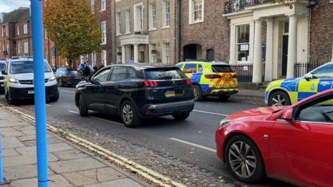 Four police cars parked on either side of Bootham, a main road into York. Traffic is trying to get past. A police officer is walking across the road.