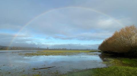 A rainbow spans this picture of a water filled landscape at Port Meadow. A tree lined horizon can just be seen in the distance with a cloud filled sky above it which is dominated by the arch of the rainbow.