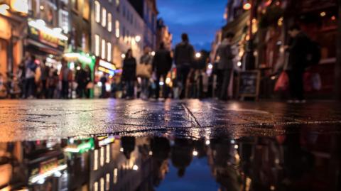 A stock image showing a busy street at night. The picture is taken from ground level with a puddle in the foreground, reflecting the people and lights from the shop windows and surrounding buildings. 