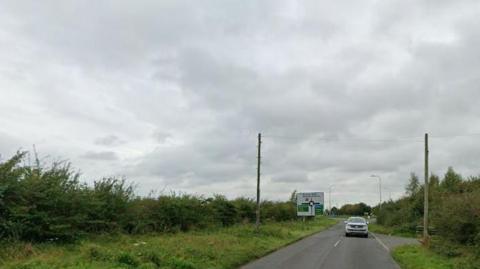A general view of Burgh Road, off which the development would be built. It is a single carriage rural road flanked by hedges.