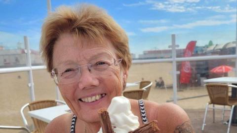 Penny Bulpitt: a woman with short brown hair wearing glasses and smiling. She is holding an ice cream cone. Behind her are tables, chairs and a beach.
