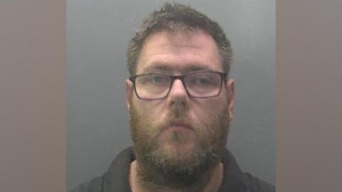 A police custody shot of Kyle Spires shows him looking at the camera. He has short brown hair, a beard and a moustache. He is wearing dark-rimmed spectacles and a dark polo top