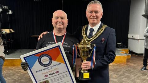 A bald man is in a black T-shirt, with black gloves and a blue, white and red medal strap around his neck. He is holding a large certificate titled "world record certificate". it reads "Paddy Doyle, 1000, 1000 fitness endurance challenges and world records". He is standing next to the Sutton Coldfield mayor, who has a suit on as well as his mayoral chains. He is holding a trophy.