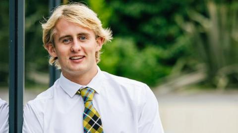 A young man with blonde hair is smiling casually. He's leaning against a window and is wearing a white shirt with a yellow and black checked tie. 