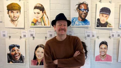 James Nickells stands in front of 8 of his portraits hanging up on a wall at his exhibition. He is wearing a brown knitted jumper and a black fedora hat.