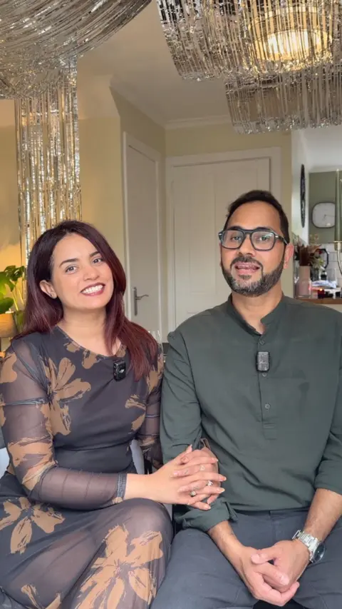 A man and a woman with their arms round each other, smiling at the camera. The woman is making a peace sign with her hand.