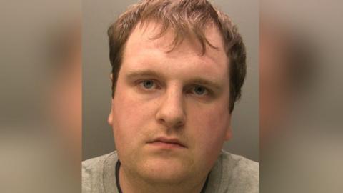 Police custody image of Jonsen Lamb. He has brown hair and is wearing a grey jumper. He is looking into the camera.