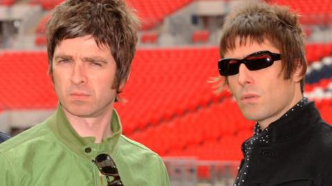 Noel Gallagher (Left) in green bomber jacket with shades hanging onto it with Liam in a black jacket and black shirt with white polka dots.