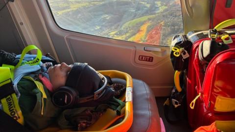 Inside a helicopter, the window in the background showing the landscape below. In the foreground, Dom, 12, lies unconscious on a stretcher with hat and coat and lots of straps to keep him in. There is a large bag of equipment to the right.