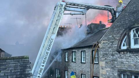 An aerial ladder with a fireman on it tackling the fire at a building in Kirkby Lonsdale. The flames and smoke are visible and a jet of water is also hitting the traditional stone building.