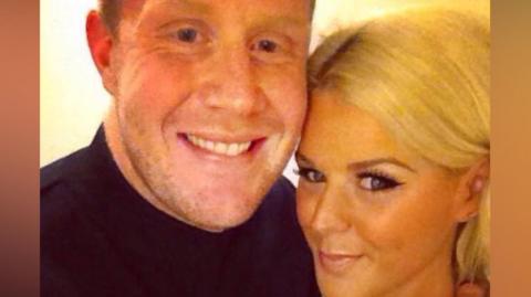 The image looks like a close‑up selfie of two people standing very close together. One person is wearing a dark top, and their arm is around the other person’s shoulder. The other person has light‑coloured hair pulled back and is wearing a sleeveless outfit with a beaded or sparkly trim along the strap. The lighting is warm, giving the whole scene a soft yellow tone.
