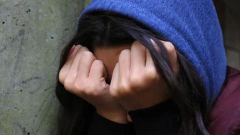 A person with long black hair with the hair in their hands, with clenched fists. They have a blue hood over their head and are next to a grey concrete wall