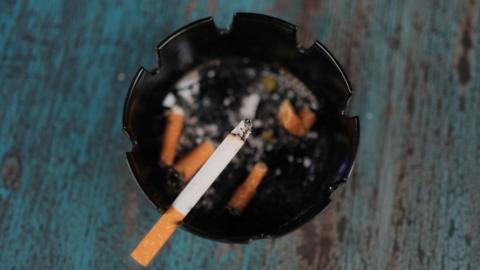 An overhead shot of a cigarette balancing on an ashtray. Several stubbed cigarettes lay crashed inside the black ashtray