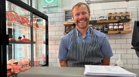 Toby wearing butcher's overalls inside his shop with meat in a glass window cabinet to his left. He is smiling at the camera