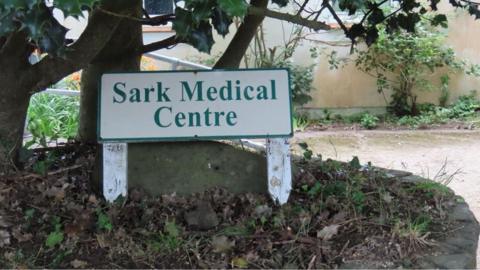 A small, white wooden sign with green writing and trim, reads Sark Medical Centre. It is in front of a holly tree and stone, on a bank of fallen leaves.