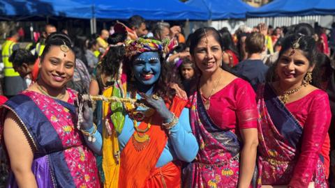 Group picture of four Hindu women smiling at the event. One of them is wearing blue facepaint and top with an orange toga draped over a shoulder as she depicts the Hindu deity Lord Krishna. She holds up a flute and wears a multi-coloured headwrap plus gold-coloured necklaces and bangles. The three ladies around her wear identical blue, pink and purple saris.
