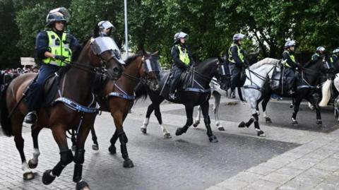 Police officers on horses. There are seven of them in total. The horses are wearing shields over their eyes and the police officers are wearing high vis jackets and helmets. 