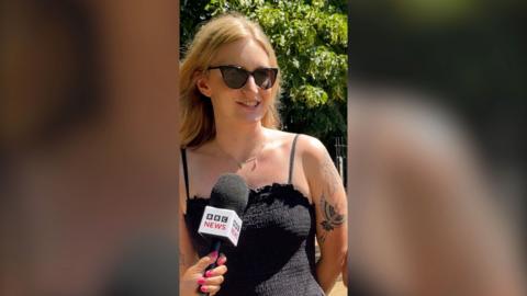 A woman speaking into a mic. She has blonde hair, sunglasses, a black strappy dress and tattoos.