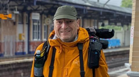 A man is wearing a bright orange jacket and is wearing a green baseball cap while standing at a railway station