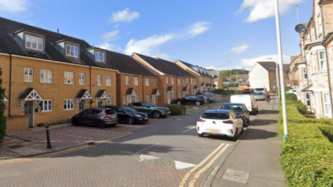 A row of brick houses with and parked cars on both sides of a narrow street.