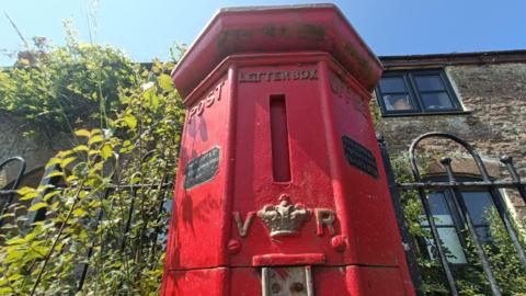 A red octagonal pillar postbox stands in front of black railing and green shrubbery. Behind can be seen a stone building with large arched windows.
