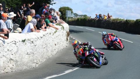 Davey Todd ahead of Dean Harrison on the road as the round a bend while spectators watch the action from behind a white painted stone wall.