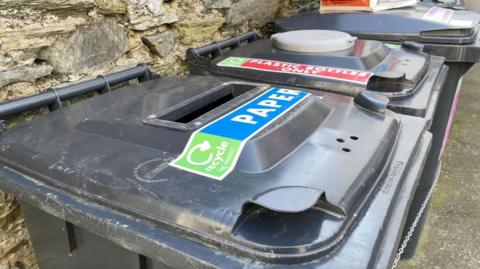 Paper recycling black bin in row of others against a wall.