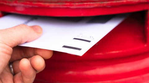 A hand holding a white letter and posting it in a red post box. It is a close-up photo of part of a post box, with the area where letters are posted being prominent in the image.
