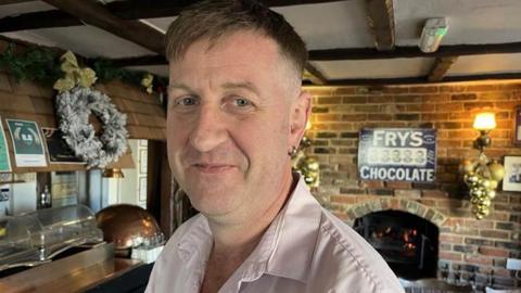 A man with grey hair and a pink shirt stands in front of a wooden pub bar and a brick fireplace. 