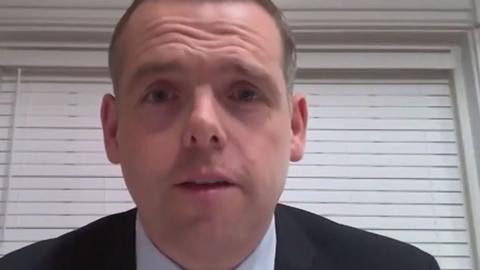 Man in black suit stands in front of a white wall looking into the camera.
