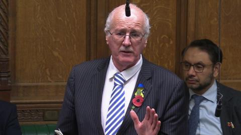 A man with white hair and glasses looks across the screen. He is wearing a navy suit with a white stripe and a blue and white striped tie. He is wearing a poppy and a pin with Ukrainian flag colours. 