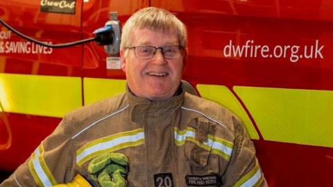Keith Prince smiling for a picture with his firefighter uniform in front of a Dorset and Wiltshire rescue fire engine.