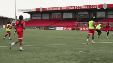 Players on a green football pitch. All are wearing red kit and some have neon green bibs on. A short stand with red seats in the background has Eastbourne Borough Football Club written across the top.