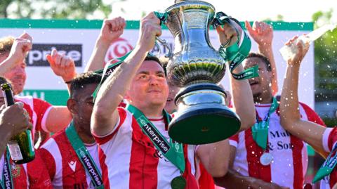 Hornchurch's Tom Wraight, in red and white striped kit, lifts the National League South play-off trophy, with his team-mates around him