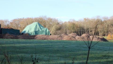 A large green tarpaulin covers a mound of materials on a field, with several piles of brown waste nearby. Trees line the background.