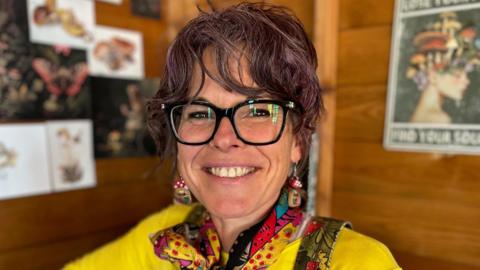 Kylie Dixon is pictured seated inside her allotment shed. She is wearing dark rimmed glasses, a yellow t-shirt and green dungarees with a brightly coloured neck-scarf and mushroom-shaped earrings. She has purple-tinted dark curly hair that is tied up.