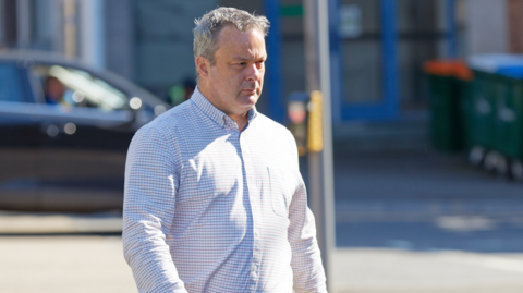 A man with grey hair walking. He has a white checked shirt on. 
