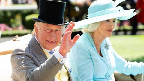 King Charles and Queen Camilla at Royal Ascot in June 2026. They are sitting in a acarraige. King Charles is wearing a top hat and Queen Camilla is in a pale blue outfit with matching hat