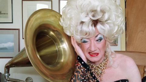 Paul O'Grady as his alter ego Lily Savage in a blonde wig next to a grammarphone 