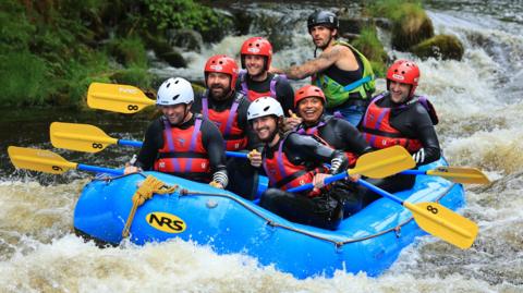 Seven men in a white water raft, each holding oars. They are all smiling, and have life jackets and helmets on, and the boat is in choppy and foaming waters