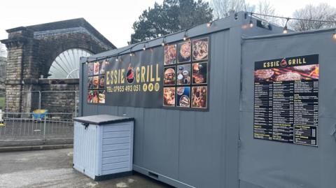 A shipping container is pictured with the words Essie Grill on a poster, with pictures of food surrounding it