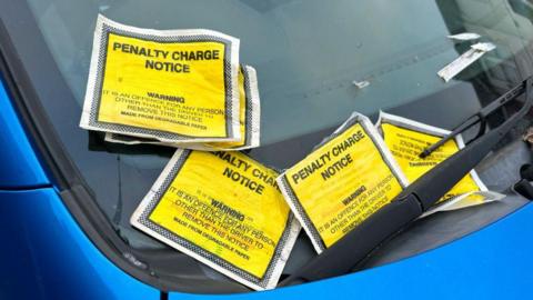 Five yellow penalty charge notices on a windscreen of a blue car. 