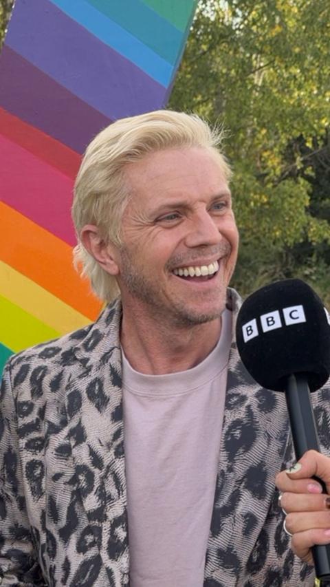Jake Shears of Scissor Sisters smiles during an outdoor interview with a BBC microphone. He is wearing a black-and-white patterned jacket over a light-coloured top. A large rainbow-coloured backdrop and green trees are visible behind him.