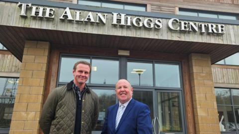 Two men are standing outside a building that has a large silver sign reading "The Alan Higgs Centre". One man has short brown hair and is wearing a brown/dark green jacket, and the other is bald and is wearing a dark blue suit jacket