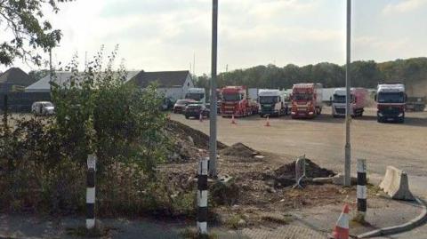 A dusty car park with a five lorries in view and some cars parked. There are trees in the background and clouds above.